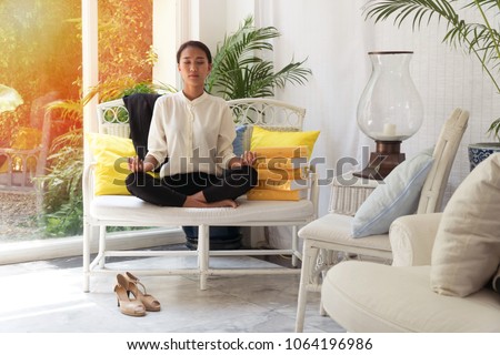Beautiful young woman meditates on a long white chair in living room with a stack of colored books left beside her in living room. Businesswoman practicing yoga at home.Relax after work concept