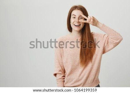 Lively adorable ginger girl showing victory or peace sign near eye while smiling broadly and expressing positive emotions over gray background. Positive coworker makes dance move