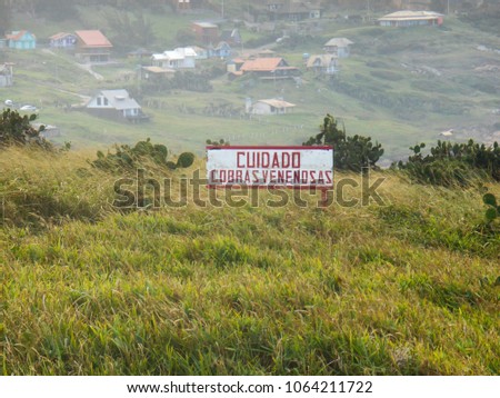 lawn with clumps of cacti and warning sign, in the background the village of Santa Marta, Brazil.  English text translation into the picture - Poisonous snake care.