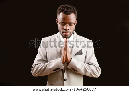 Close up portrait of a serious young smart Afro man with joined hands and closed eyes on the isolated black background