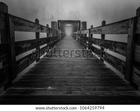 A haunting black and white image of an old truss footbridge over the lake on a foggy morning at Tims Ford State Park in Winchester Tennessee. Resembles a portal in a sci-fi movie.