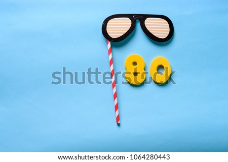 80 celebrating yellow number with sunglasses cute paper mask on straw stick . Eighty Modern alphabet digits on blue background. 80 th birthday party anniversary card.Flat lay, top view.