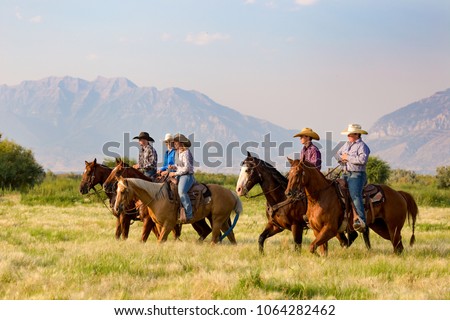 A group of five young people horseback riding in the grassy fields with the mountains of Utah in background