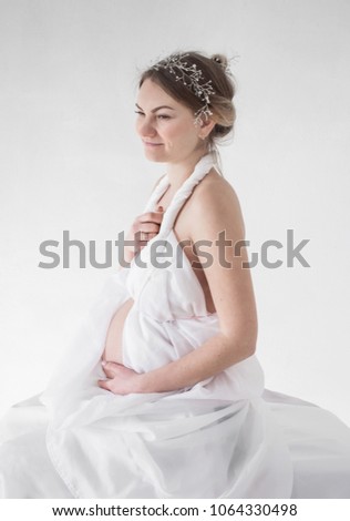 photo portrait of a pregnant girl in a diadem in a white drapery on a white background sits in a profile