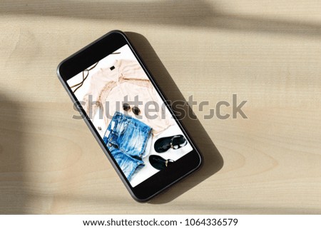 Online shopping concept. Smartphone with photo Summer set of female clothes. Female denim shorts with blouse and sandals on the mobile phone screen, top view, flat lay