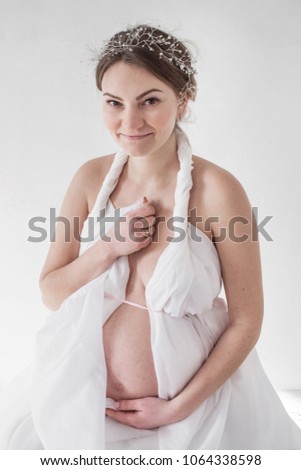 photo portrait of a pregnant girl in a diadem in a white drapery on a white background sits