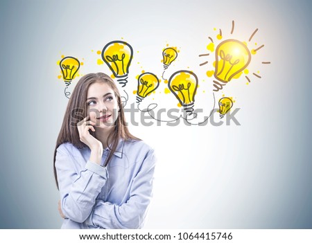 Dreamy young woman with brown hair wearing a blue shirt and business suit pants is looking upwards and thinking. Yellow light bulbs on a gray wall. An idea concept