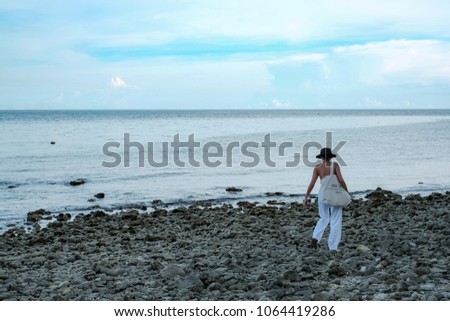 Asian girl tourist in vacation wearing black hat and cotton bag walking on stone beach with beautiful sea and sky background, asian woman holiday portrait on rock beach showing relax time and freedom
