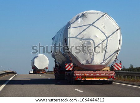 Two large lowbed semi trucks with a police escort car carrying oversized cargo big white barrels drive on the asphalt highway road at summer day against blue sky background, close up back view Royalty-Free Stock Photo #1064444312