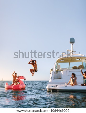 Young man jumping off the boat in to the sea. Young people having fun during party on a private boat. Men and women on yacht and inflatable toy in sea. Royalty-Free Stock Photo #1064469002