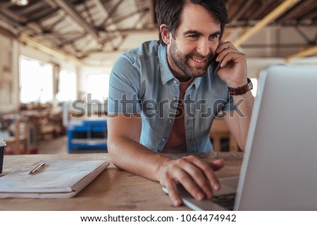 Smiling young craftsman standing at a workbench in his large workshop working online with a laptop and discussing designs with a customer on a cellphone Royalty-Free Stock Photo #1064474942