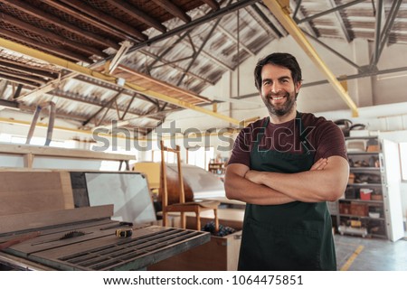 Portrait of a smiling young woodworker standing with his arms crossed by a bench saw in his workshop full of carpentry equipment Royalty-Free Stock Photo #1064475851