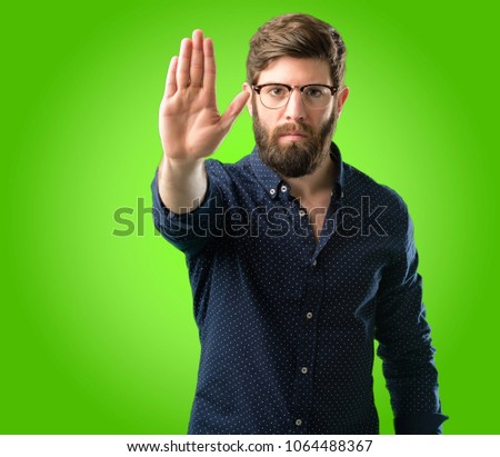 Young hipster man with big beard annoyed with bad attitude making stop sign with hand, saying no, expressing security, defense or restriction, maybe pushing over green background