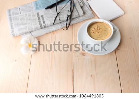 Coffee cup on table, plumeria flower, blurred reading glasses and pen on newspaper, top view. Good morning coffee.