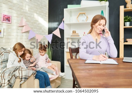 Smiling young woman taking notes while discussing project details with client, her little daughters wrapped up in computer game while sitting on cozy sofa of living room