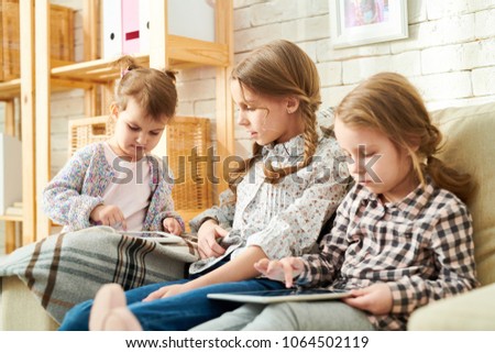 Three little sisters gathered together at cozy living room illuminated with sunbeams and playing computer games on digital tablets