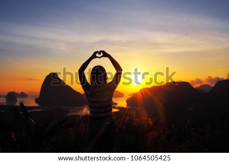 Silhouette picture of lady doing hand sign in heart shape on the top of the mountain with beautiful landscape view of islands in andaman ocean and sunrise sky in the morning on background