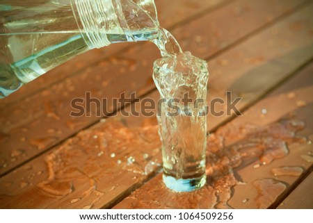 The water is poured from the jug into a glass and poured onto a wet wooden table with drops or dew.