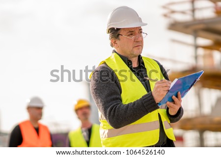 Foreman at work on construction site checking his notes and drawing plan on clipboard Royalty-Free Stock Photo #1064526140