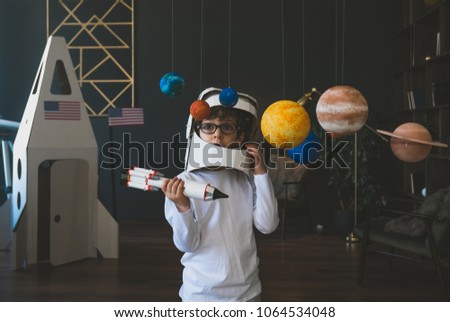 Cute little boy wearing cardboard astronaut helmet flying toy rocket through planets, cardboard spaceship rocket in the background Royalty-Free Stock Photo #1064534048
