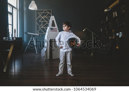 Cute little dreamer boy posing with a helmet at home, pretending to be an astrounaut, cardboard space rocket in the background Royalty-Free Stock Photo #1064534069