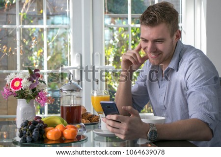 Handsome man sitting at breakfast table with cereal, fruit, flowers, coffee and croissants using his mobile device phone and smiling laughing 