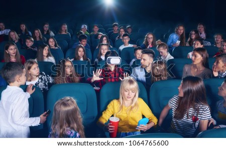 Cinema hall full of young people and children,looking surprised and exited, watching on girl wearing 3D-eyeglasses. People sitting on comfortable plases feeling interested.