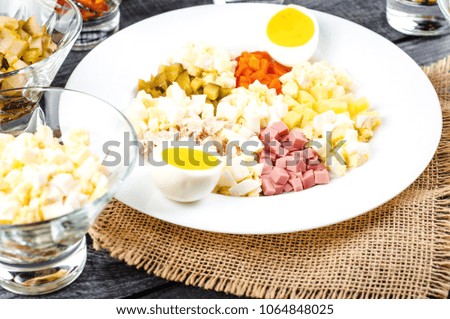 Ingredients shredded for salad in transparent plates on a black wooden background. Olives, pickled cucumbers, sausages, carrots, pickled green peas, boiled eggs and potatoes, white sauce, parsley.