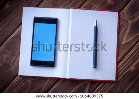 A modern digital smartphone with a bright screen lying on a books desk with an open paper pad and a pen