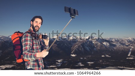 Hipster carrying backpack with camera and taking selfie while standing against mountains