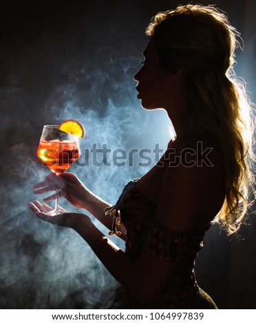 Beautiful young woman profile in backlight drinking aperol spritz isolated black background with smoke. Club party menu concept