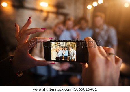 Close up focus view of woman hands and mobile while taking a photo of four happy friends relaxing in the pub.