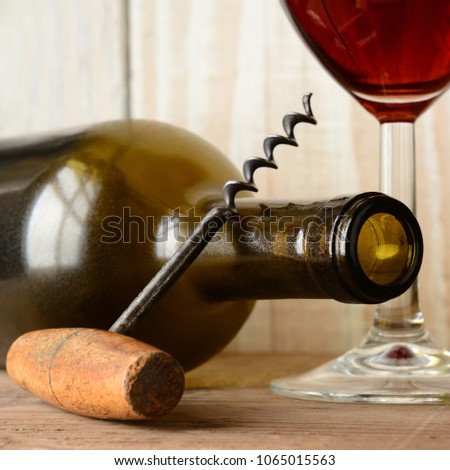Corkscrew leaning on an empty bottle of red wine with full glass behind. Closeup in square format.
