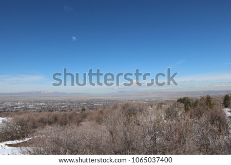 A view from the Bountiful Hills with snow, scrub oak trees and bushes, pine trees, blue sky with clouds, and Farmington Bay and Utah mountains in the distance