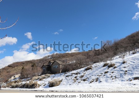 A mountain home,  with trees, sagebrush and bushes, a rock retaining wall, snow, and a blue sky with clouds in Utah