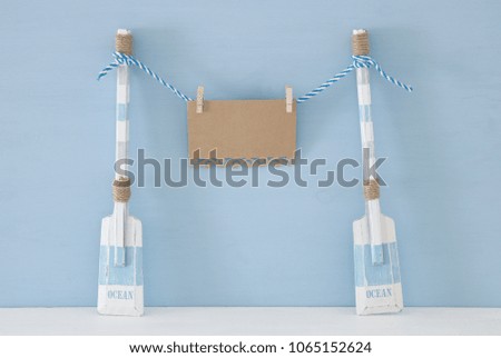 nautical concept with wooden decorative boat oars and empty note for copy space hanging on a string over blue background