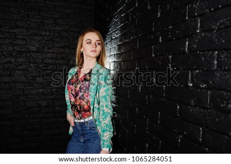 Stylish blonde girl in jacket and jeans against brick black wall at studio.