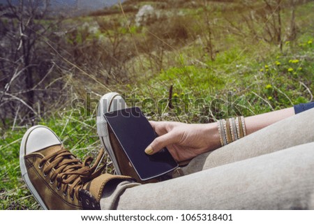 Woman Sitting Outside In Nature On Vibrant Grass In A Sunny Day Holding Black Smartphone With Empty Screen In Hands 