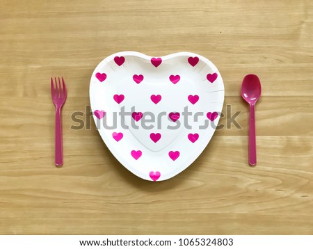 Closeup A pink heart printed paper plate with plastic spoon & fork on wooden background.The concept of Love,Valentine day,wedding celebration,anniversary party,food display.Selective focus.Copy space.