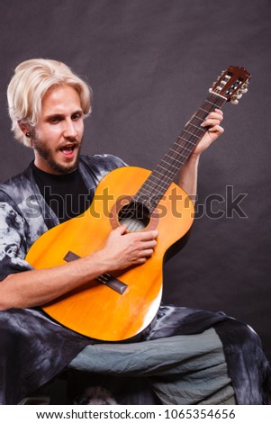 Music, passion concept. Young blonde man wearing fancy shirt playing on acoustic guitar and singing, studio shot, black background.
