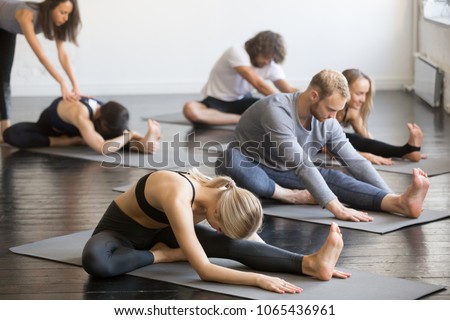 Group of young sporty people practicing yoga lesson with instructor, doing Janu Sirsasana pose, Head to Knee Forward Bend exercise, working out, in yoga studio Royalty-Free Stock Photo #1065436961