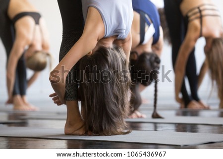 Group of young sporty people practicing yoga lesson, standing forward bend, head to knees exercise, uttanasana pose, working out, indoor close up view photo, studio