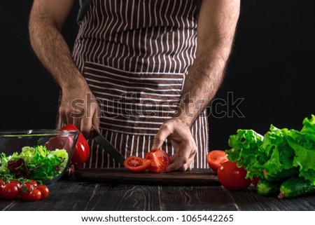 Man preparing salad on a wooden table. Men's hands cut the tomato to make a salad on black background