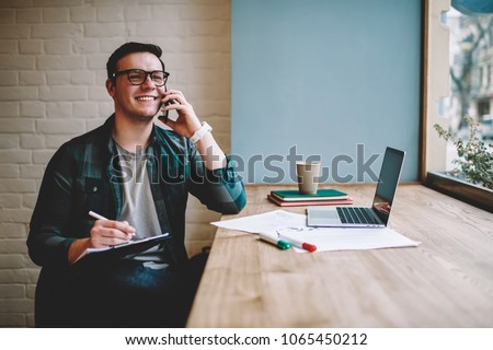 Cheerful male freelancer making telephone call share good news about project working in cafe interior,happy hipster guy having smartphone conversation while studying in good mood writing in planner Royalty-Free Stock Photo #1065450212