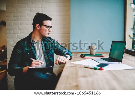 Pensive hipster guy in eyewear watching training webinar on laptop computer spending time on e learning, male designer working on freelance browsing information for project on netbook drawing sketch Royalty-Free Stock Photo #1065451166