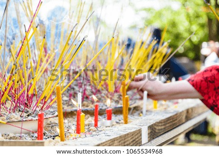 Close-up view of burning incense sticks planted into piles of sand on a table, diffusing a bluish smoke during a buddhist ceremony, with candles in the foreground and female hand in the background.