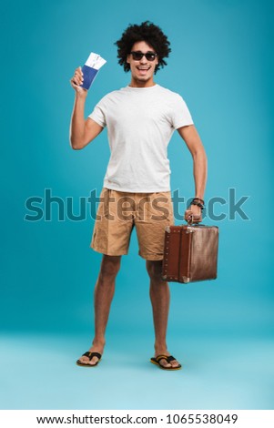 Picture of handsome happy young african curly man tourist isolated over blue wall background holding tickets with passport and suitcase.