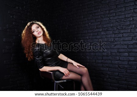 Brunette curly haired long legs girl in black leather dress posed at studio on chair against dark brick wall.