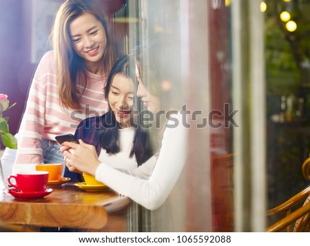 three happy beautiful young asian women sitting at table chatting talking playing with cellphone in coffee shop or tea house, shot through window glass.