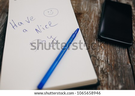 White notebook with a blue pen is lying on the old vintage background. Have a nice day wish is written on sketch book. Black smartphone with earphones is lying nearby. Modern technologies.